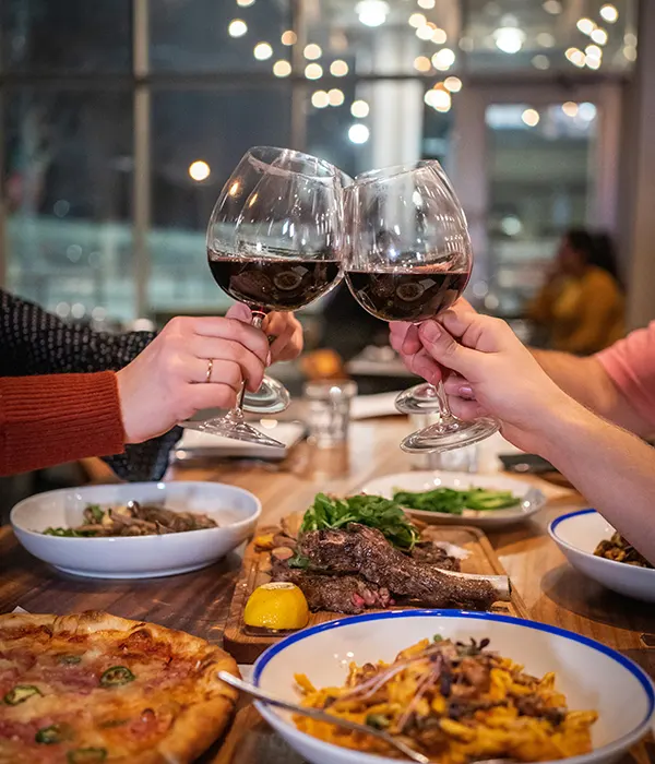 Ein Tisch mit italienischem Essen, darunter Pizza, Steak, Pasta und grünes Gemüse. Es gibt vier Hände auf dem Foto, die alle Weingläser mit rotem Wein halten. Die Personen erheben ihre Gläser, um sie zu stoßen.